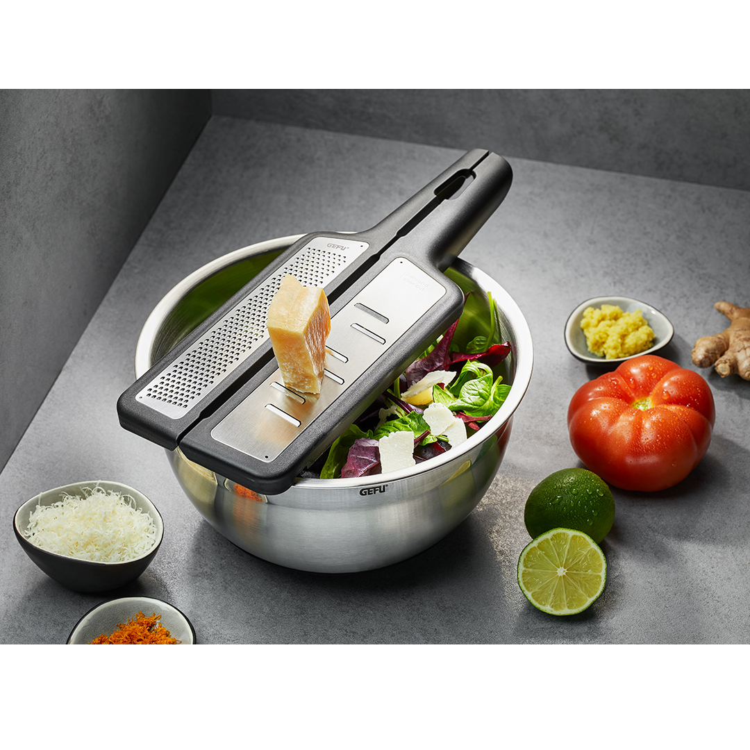 Uz pelēka galda novietota bļoda ar svaigiem salātiem un  divdaļīgu rīvi ar cieto sieru uz tās